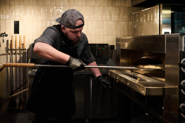 Young male chef in uniform putting pizza into hot electric oven for baking while standing in front...