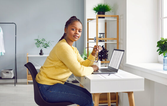 Young woman works remotely and uses electronic business sheets. Beautiful African American girl sitting at desk with laptop computer at home, holding glasses and looking at camera. Side, profile view