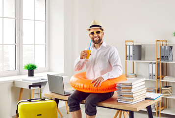 Funny office worker ready to go on summer holiday. Happy smiling man in white shirt, sun hat beach ring and sunglasses drinks orange juice, sitting on desk with laptop and papers. Annual leave concept - obrazy, fototapety, plakaty