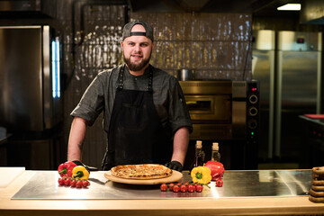 Happy young successful chef and owner of restaurant or pizzeria looking at camera while standing by table with appetizing pizza in the kitchen - obrazy, fototapety, plakaty
