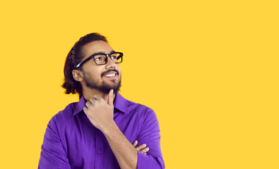 Fototapeta na wymiar Happy Indian young man with thoughtful expression thinking about plan or having idea in mind. Man in glasses and shirt is smiling and holding his chin while looking at copy space on orange background.