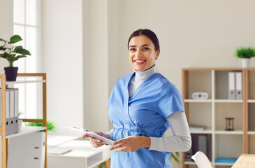 Fototapeta premium Portrait of a young pretty happy smiling female doctor in blue medical uniform standing in her office. Confident woman physician holding report file with appointment and looking at the camera.