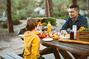 Father dad and school kid boy child having a picnic in the forest camping site with vegetables, juice, coffee, and croissants. Wooden crate with fresh organic veggies surrounded with bread baguettes