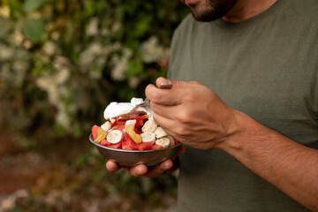 Cropped photo of young man holding tropical fruit salad with cut various exotic fruits metal bowl, fork, eating outside.