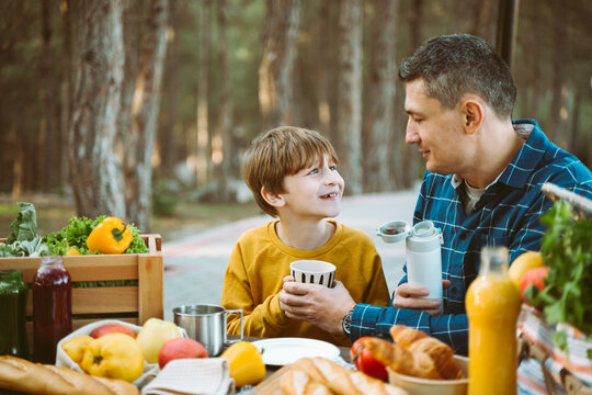 Father dad and school kid boy child having a picnic in the forest camping site with vegetables, juice, coffee, and croissants. Wooden crate with fresh organic veggies surrounded with bread baguettes