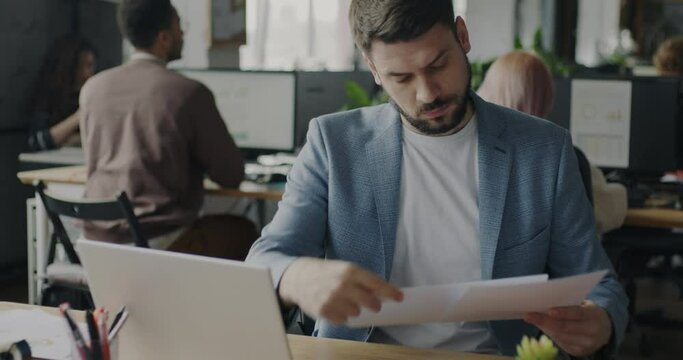 Portrait of male specialist reading business documents and working with laptop in open space office. People and corporate work concept.