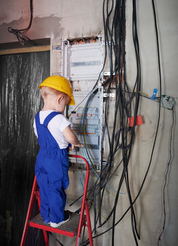 Back view of child electrician standing on ladder near electrical switchboard in apartment under renovation. Kid in safety helmet and work overalls repairing electric control panel at home.