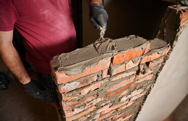 Close up of male hands in work gloves laying brickwork in building under construction. Man bricklayer builder applying cement mortar on bricks with trowel tool. Masonry construction concept.