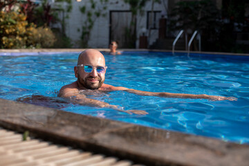 Portrait of young bearded man swim in swimming pool, relaxing, cooling freshening up, looking at camera on sunny day.