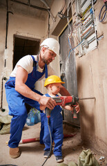 Man and little boy construction workers drilling wall with electric drill in apartment under...