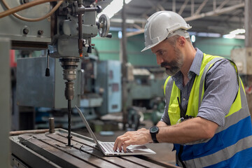 Caucasian man engineer wear white hard hat and wearing safety uniform using laptop working steel drill machine at heavy industrial factory. Male working machine lathe metal.