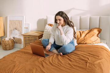 young curly girl in home casual clothes studying online at home sitting on the bed in her bedroom