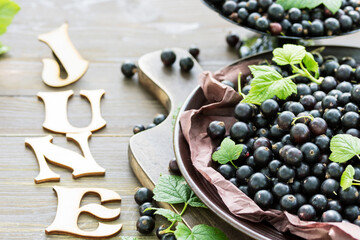 Freshly gathered big juicy black currants on brown plate closeup, black currant harvest, word July of wooden letters on table, berries outdoors, in garden