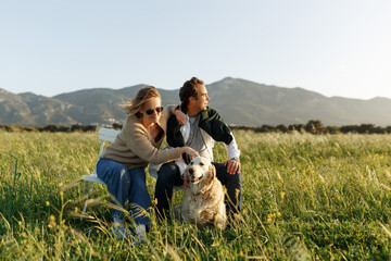 Young happy couple with dog, sitting on chairs in feather grass field on summer sunset day