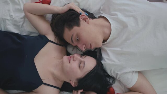 Loving young couple lying down on white bed smiling caressing top view. Close-up portrait of happy confident Caucasian man and woman resting at home indoors