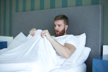 Young happy man looking at his penis under blanket, surprised guy is having good morning potency, erection in bed in bedroom  - obrazy, fototapety, plakaty