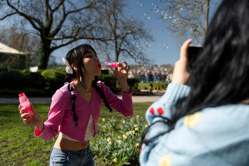 Fototapeta premium Young woman recording friend blowing bubbles in park