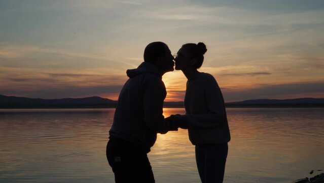 A couple in love stands on the shore of the lake at sunset. Hugs and kisses gently. Feeling, love and warmth.