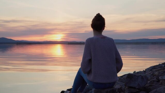 The girl sits on the shore of the lake at sunset, admiring the beauty of nature. Waving his arms to scare away mosquitoes and midges.