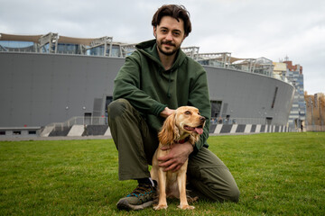 Smiling man posing together with his dog outdoors. A bearded brunette man sits on the grass and...