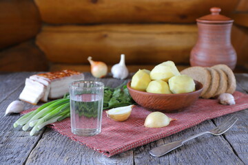 On the wooden table against the backdrop of a log wall is a glass of vodka next to vegetables, greens, bacon, bread and boiled potatoes in a clay bowl.