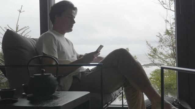 Backlit Man Sits in Sofa Checking his Smartphone, Ocean View in Window