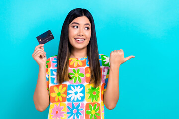 Photo of cheerful dreamy lady wear flower print t-shirt smiling holding credit card thumb empty space isolated blue color background