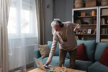 Happy senior old grandfather man in wireless headphones dancing, singing at home, choosing favorite...