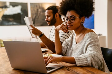 Smiling african woman working on laptop while her husband reading sitting at table