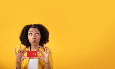 Dreamy black lady with credit card enjoying online shopping, looking at free space isolated on yellow studio background