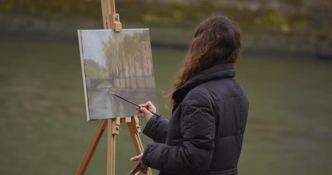 Woman painting a picture near the river Seine. Creative woman artist paints a picture with a brush while standing near the easel in Paris. Woman artist, art for sales, inspiration