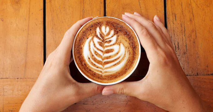 Woman's hands take cup of coffee and raise it above the table, rosetta image is drawn on the foam. Tasty drink with subtle and short-living art made on milk froth