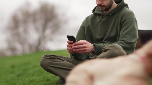 A man sits on the grass with a phone in his hands and puppies run around him. A brunette man is relaxing outdoors in the park with his dogs.
