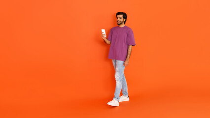 Fototapeta na wymiar Positive young indian guy walking with smartphone on orange