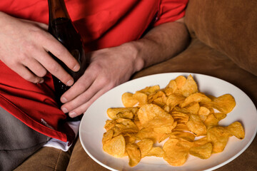 A football fan in a red jersey is watching a match on TV, sitting on a couch, drinking beer, and eating chips. Close-up, unrecognizable, no face.