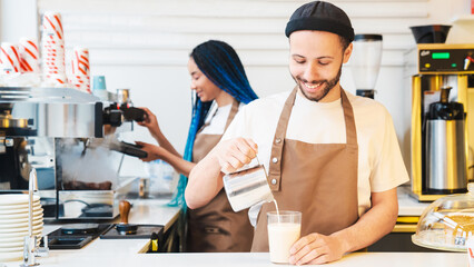 Happy male barista pouring milk into the glass with coffee while making latte art in coffee shop