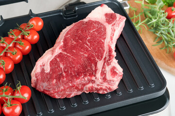 Raw prime steaks on electric grill. Black angus prime meat steaks Ribeye on modern electric grill with rosemary, cherry tomato and pepper. Cooking process in modern kitchen. Top view, copy space.
