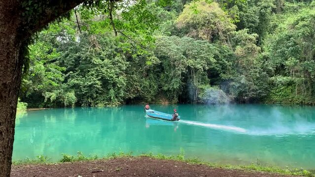 Small yola fishing boat leaving a magical blue lagoon surrounded by trees in the Caribbean and disappears through the trees leaving a trail of engine smoke