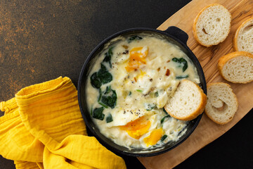 Healthy breakfasts or dinners, fried eggs with cheese, spinach leaves and coconut milk in a cast-iron pan, sliced baguette on a cutting board on a brown background