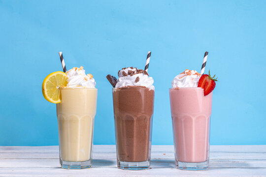 Set of summer refreshing milkshakes or smoothies with classic delicious taste - vanilla lemon, strawberry, chocolate, with whipped cream and toppings, On a white wooden light blue sunny background 