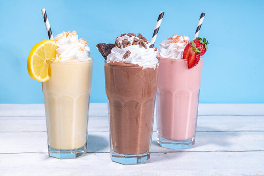 Set of summer refreshing milkshakes or smoothies with classic delicious taste - vanilla lemon, strawberry, chocolate, with whipped cream and toppings, On a white wooden light blue sunny background 