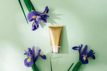 Golden cosmetic cream tube and blue iris flowers on light green background. Top view, flat lay,...