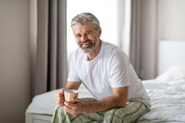 Happy middle aged man sitting on bed, drinking coffee