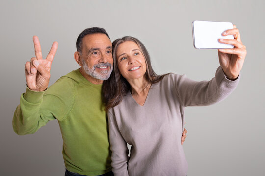 Smiling caucasian elderly man and lady pointing peace sign with fingers and taking selfie on smartphone