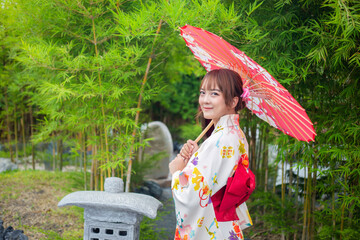 Young woman wearing traditional Japanese kimono or yukata holding an umbrella in a park with bamboo...