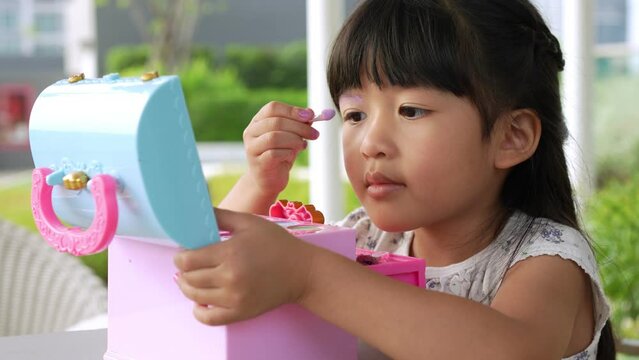 Adorable little child Asian girl paints her eyes with pink children heads and looks in the mirror. A child plays at home in a toy beauty salon. Increase learning development for preschoolers.