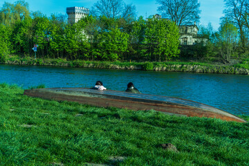 Two people are sitting next to a wooden boat on the river bank with a vintage building among the trees in the background, environmental art, sunny spring day, relaxing in unity with nature