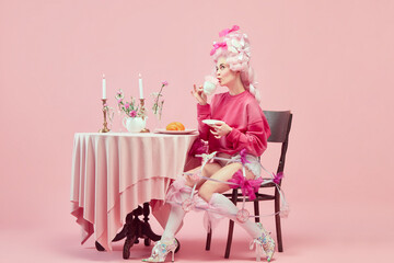 Portrait of elegant princess, queen wearing pink clothes, and wig drinking coffee over pink...