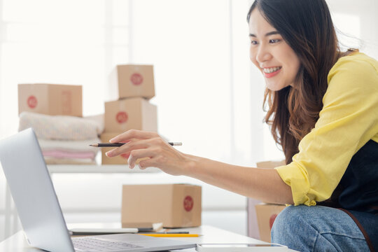 Asian woman packing parcel boxes and checking orders from laptop, she owns an online store, she packs and ships through a private transport company. Online selling and online shopping concepts.