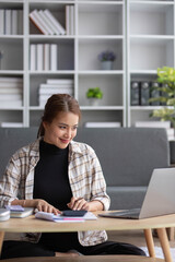 Attractive Asian female freelancer planning her project, working on her tasks on laptop computer at a coffee table in her living room.